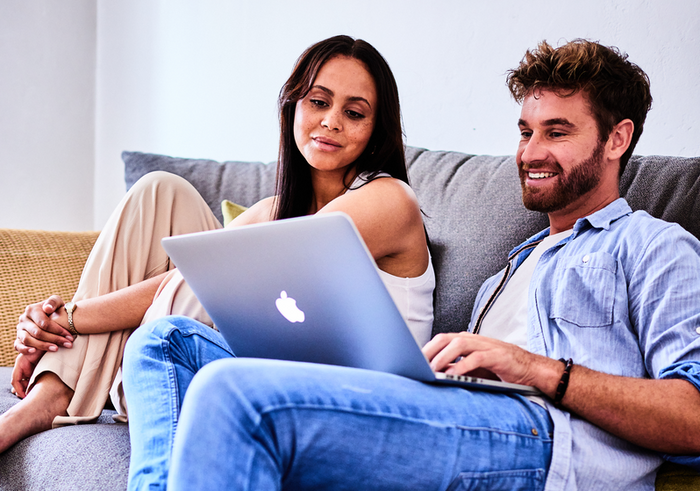 Junges Paar sitzt entspannt auf einem Sofa und schaut gemeinsam auf einen Laptop.