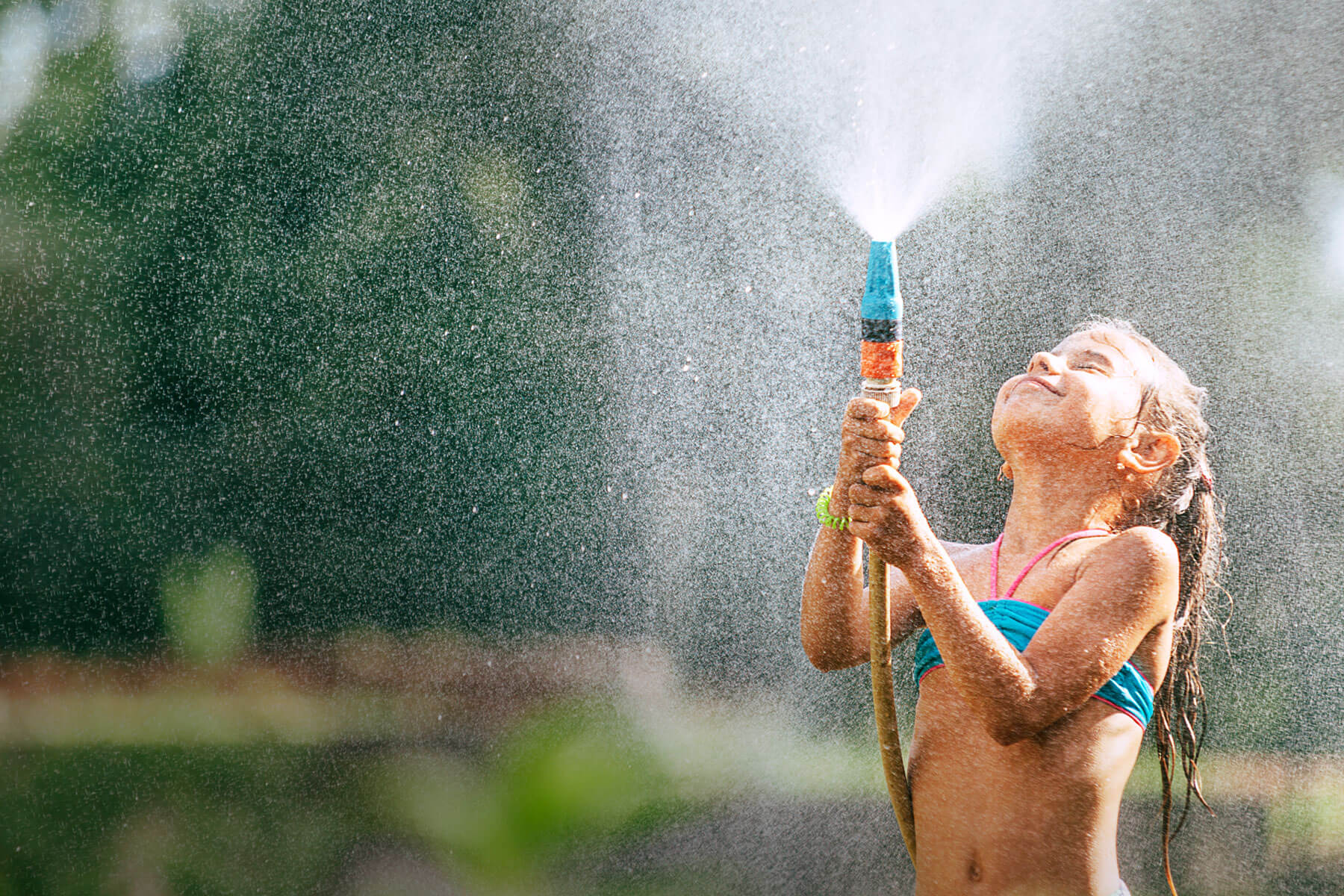 Bild eines kleinen Mädchens, dass im Garten mit einem Wasserschlauch Wasser in die Luft spritzt und dabei nass wird.